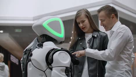 A-man-and-a-woman-in-the-Mall-interact-with-a-robot-consultant-by-tapping-the-screen-and-smiling.-Cyborg-Android-helps-people
